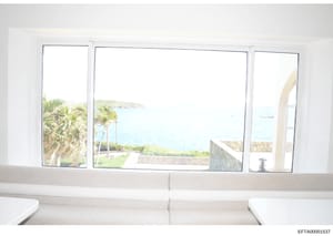 This is a photograph showing the interior of a waterfront property, taken from inside a room looking out through large floor-to-ceiling windows. The image captures a scenic coastal view with palm trees, ocean water, and distant shoreline, and appears