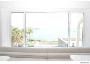 This is a photograph of a luxurious interior space with expansive windows offering panoramic views of a tropical coastal landscape. The image shows a modern residential or commercial property with floor-to-ceiling windows framing palm trees, ocean wa