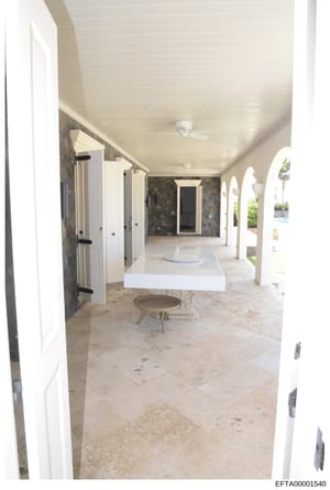 This is a photograph of an upscale residential interior space, showing a covered outdoor or semi-outdoor area with white columns, stone flooring, white cabinetry, and modern finishes. The space appears to be a loggia, pavilion, or pool house area fea