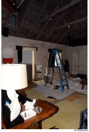This photograph shows an interior room in active renovation or construction, featuring exposed wooden ceiling beams, protective coverings on the floor, a blue ladder, and various construction materials. The space appears to be a residential property 