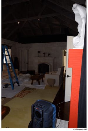 This photograph shows an interior room in the process of renovation or construction, featuring a fireplace, exposed structural ceiling elements, and various construction equipment and materials. The image appears to be evidence documentation from a p