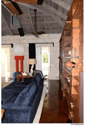 This is an evidence photograph showing the interior of a residential room, likely from one of Epstein's properties. The image depicts a bedroom or living space with contemporary furnishings including a blue velvet sofa, wooden shelving, exposed ceili