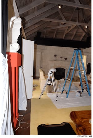This photograph shows an interior space under active renovation or construction, featuring exposed wooden ceiling beams, ladders, protective coverings, and ongoing work. The room appears to be a large space with high ceilings and a fireplace, suggest