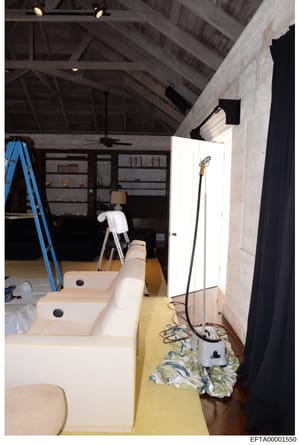 This photograph documents an interior space during renovation or construction, showing built-in seating with cream-colored cushions, exposed ceiling beams, shelving units, and various construction materials. The image appears to be from a property in