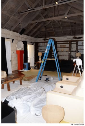 This photograph shows a spacious interior room undergoing renovation or construction, featuring exposed beam ceilings, protective plastic sheeting, a blue ladder, and various furniture and decorative items. The image documents the interior condition 
