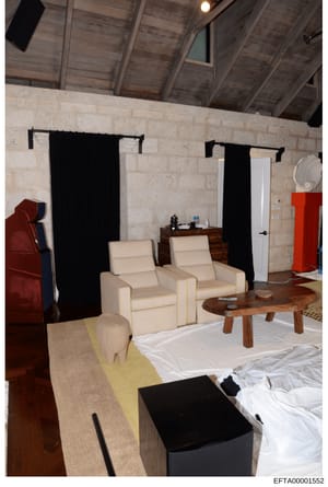 This photograph depicts an interior room with modern luxury furnishings, featuring travertine stone walls, exposed wooden beam ceiling, cream-colored lounge chairs, wooden accent tables, and black drapery panels. The space appears to be a high-end re