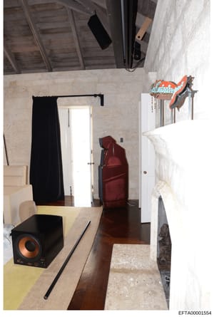 This photograph shows an interior room, likely a bedroom or storage area, featuring travertine or marble-textured walls, wooden flooring, and various items including a red luggage bag, black speaker with orange accents, decorative bird artwork, and b