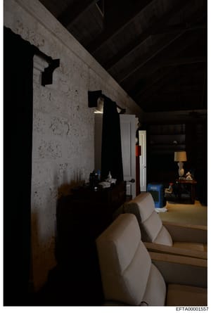 This photograph documents an interior space, likely a basement or lower level room within a property under investigation. The image shows a stark, sparsely furnished room with exposed beam ceiling, concrete/plaster walls showing age and stains, white