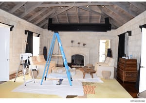 This photograph documents an interior space undergoing renovation or construction. The room features exposed wooden beam ceiling, a fireplace with dark surround, limestone or stone walls, and multiple doorways. Construction equipment including a blue