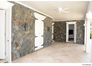 This is a photograph of a large interior room with distinctive slate or stone walls featuring earth tones and rust-colored patterns. The space shows multiple white doors with black hardware, cream-colored ceiling with ceiling fan, and light tile floo