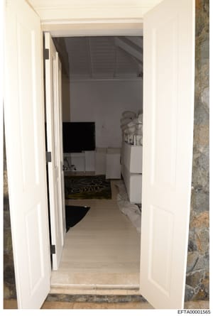 This is a photograph showing the view through an open white double door into what appears to be a bedroom or living space. The image captures the interior layout of a room with furnishings, a television, and architectural details including exposed ce