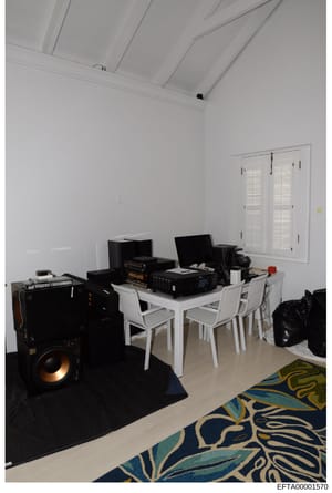 This is a photograph of an interior room containing electronic equipment, audio speakers, computer workstations, and furniture. The room features white walls, a vaulted ceiling with recessed lighting, white plantation shutters, and decorative rugs. T