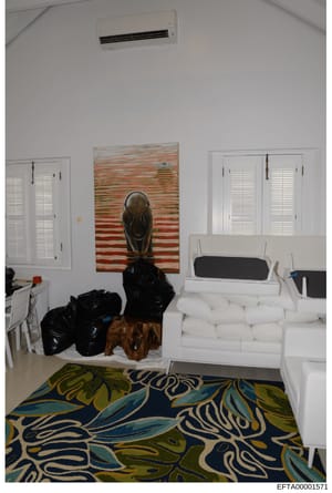 This photograph shows an interior residential space with white furniture, decorative artwork, and various items. The room appears to be a bedroom or living area with a bed, closets, air conditioning unit, and contemporary artwork on the walls. The im
