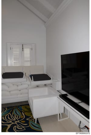 This is a photograph of a contemporary living room interior featuring white modular furniture, including a sectional sofa with black cushions, white side tables, and entertainment setup with a large black television. The room has white walls, vaulted
