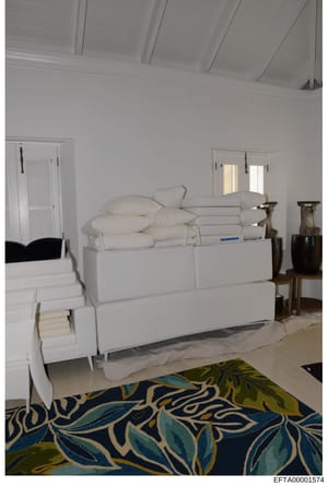 This photograph shows the interior of a bedroom featuring a white upholstered bed with pillows and folded linens, white shuttered windows, decorative wooden urns, and a patterned area rug in blue, green, and cream tones. The high white ceiling and mi