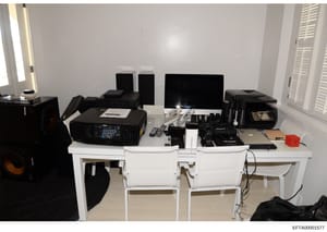 This is an evidence photograph showing an interior office space containing computer workstations, audio/visual equipment, and multiple electronic devices. The room features white walls, shuttered windows, and professional-grade audio equipment. The p