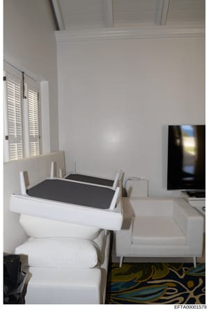 This is a photograph of an interior room showing a toilet converted into a seat or bench with a black cushioned top, positioned next to a white sofa and television. The room features white walls, shuttered windows, high ceilings, and a decorative are