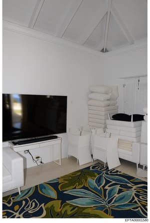 This is a photograph of a modern bedroom interior featuring white furniture, stacked linens, a large flat-screen television, and contemporary décor. The image appears to be documentation of a property's interior, likely taken during an investigation 