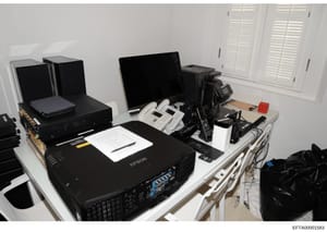 This photograph documents a computer workstation and office equipment setup, featuring multiple monitors, an Epson projector, networking equipment, white shuttered windows, and various electronic devices arranged on tables. The image appears to be ev