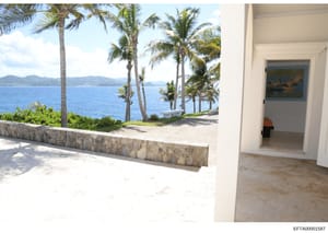 This photograph shows a beachfront or oceanfront property featuring a modern patio or deck area with direct views of the ocean, palm trees, and distant mountains. The image captures both interior and exterior spaces, including what appears to be a do
