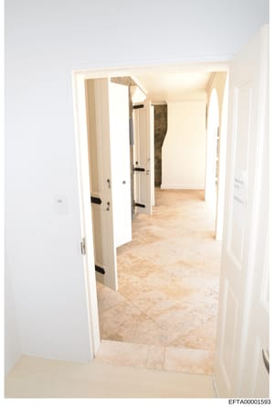 This is a photograph of an interior hallway or corridor, showing white-painted doors with black hardware, beige/cream colored walls, and light travertine or marble flooring. The image appears to be documentation of a property interior, likely taken d