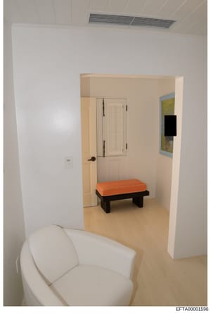 This is a photograph of an interior room showing modern, minimalist design. The space features white walls, beige flooring, a white curved chair in the foreground, a black bench with orange cushion, and a closet with louvered doors. A map or artwork 