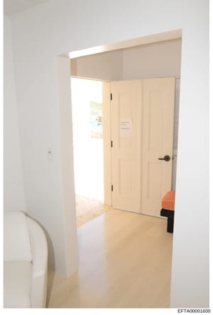 This is a photograph of an interior room showing a bedroom or living area with light wood flooring, white walls, cream-colored closet doors, and modern lighting fixtures. The image appears to be evidence documentation from a property associated with 