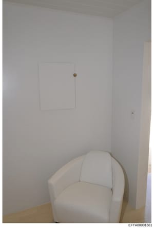This is a photograph of an interior room space showing minimalist décor. The image depicts a white-walled room with a cream-colored upholstered armchair in the foreground and a rectangular wall-mounted panel or artwork on the wall. The photo appears 