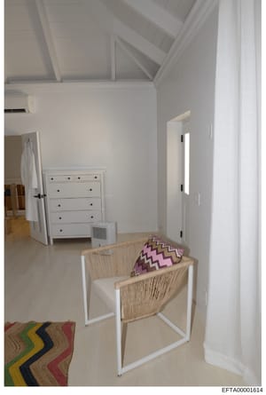 This is a photograph of a bright, minimalist bedroom interior featuring white walls, vaulted ceilings, and contemporary furnishings. The room contains a woven accent chair with decorative pillows, a white dresser, and geometric patterned area rug. Th