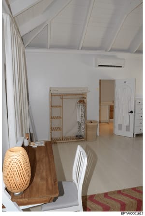 This is a photograph of a bright, minimalist bedroom interior featuring white walls, exposed beam vaulted ceiling, natural wood furnishings, and sparse decor. The room contains basic bedroom furniture including a wooden clothing rack, woven storage b