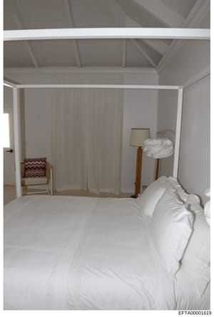 This is a photograph of an interior bedroom space, showing a four-poster bed with white linens, sheer white curtains, wooden furniture including a chair with burgundy patterned cushion, a floor lamp with beige shade, and exposed beam ceiling details.