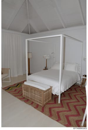 This photograph shows a modern, minimalist bedroom interior featuring a white four-poster bed with white linens, light wood furnishings, and a geometric patterned rug in red and natural tones. The room has high vaulted ceilings and appears to be part