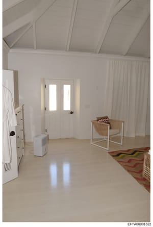 This photograph shows the interior of a sparsely furnished room with modern, minimalist design. The space features white walls, vaulted ceilings with exposed beams, polished light-colored flooring, and minimal furnishings including a wooden chair, sm