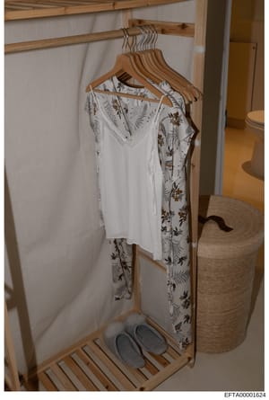 This is a photograph of a wooden clothing rack with hangers holding garments, located in what appears to be a bedroom or dressing area. The image shows a white garment with floral patterns (ferns and botanical designs in brown/tan tones) hanging on w