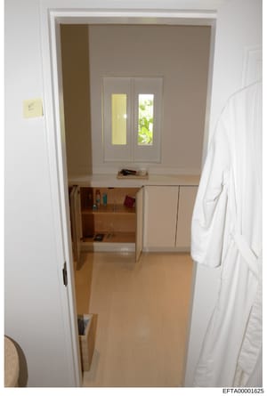 This is an evidence photograph showing the interior of a room at an Epstein property. The image depicts a bedroom or dressing area with taupe-colored walls, a wooden desk with storage, white double doors with glass panels opening to an exterior area,