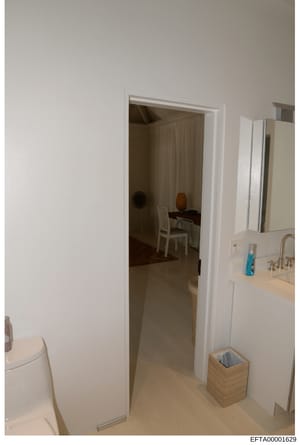 This photograph shows the interior of a residential bathroom with an open doorway revealing a bedroom beyond. The image appears to be evidence documentation from a property inspection or seizure, showing basic furnishings and room layout. The Bates n