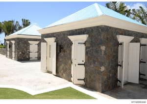 This is a photograph of a distinctive stone structure featuring multiple individual entrance doors, a bright blue metal roof, and cream-colored door frames. The building appears to be a changing room, bathhouse, or similar facility located on a tropi