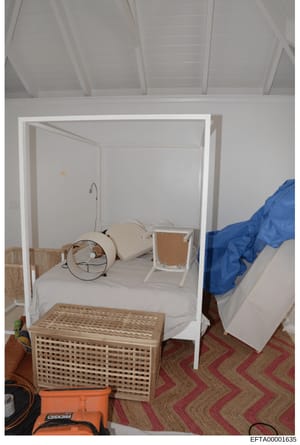 This photograph shows a bedroom interior featuring a white canopy bed frame with minimal bedding, a white wooden nightstand, and various other furnishings. The room appears to be part of a property inventory or evidence documentation from the investi