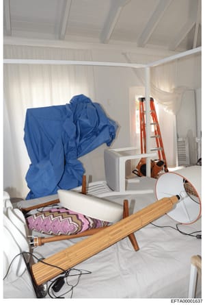 This photograph shows an interior space undergoing renovation or construction, with furniture and equipment covered with protective sheeting. The room features white walls, exposed ceiling beams, and contains various items including a blue dust cover
