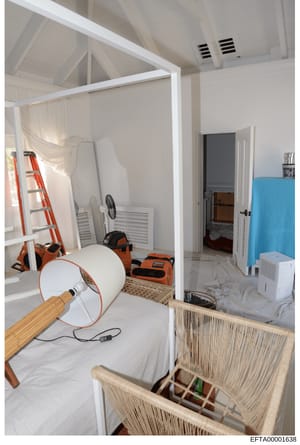This photograph documents the interior of a room that appears to be undergoing renovation or construction work. The space contains various work equipment including ladders, fans, cleaning supplies, and power tools. The room features high ceilings wit