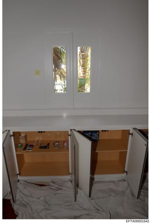 This is an evidence photograph showing a residential interior space with white cabinetry, glass doors with views to tropical landscaping, and wooden storage cabinets below. A yellow evidence marker labeled 'F' is visible on the wall. The photo appear