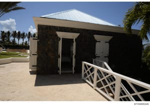 This is a photograph of a modern, luxury poolside structure featuring dark stone walls, white trim, and a metal roof. The building appears to be a cabana or changing facility adjacent to a swimming pool on an island property. The architectural style 