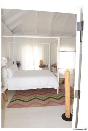 This is a photograph of a modern, minimalist bedroom interior featuring a white four-poster canopy bed, geometric patterned area rug, and clean, light aesthetic with white walls and vaulted ceilings. The image appears to be an architectural or interi
