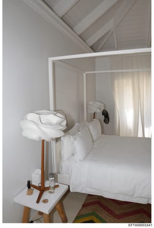This is a photograph of a minimalist bedroom interior, featuring a white four-poster bed with white linens, a wooden nightstand with a glass of water, and vaulted ceiling with striped detailing. The image appears to be architectural or interior desig