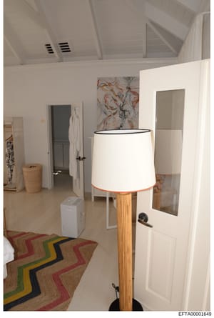 This is a photograph of a contemporary interior space showing a bedroom or guest suite with modern furnishings and design elements. The room features white walls, vaulted ceilings with exposed beams, a wooden floor lamp, abstract artwork, and access 