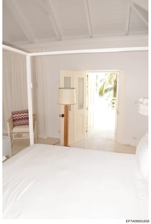 This is a photograph of a luxurious bedroom interior featuring all-white décor, likely from one of Jeffrey Epstein's properties. The image shows a spacious master suite with high ceilings, floor-to-door windows with views of tropical foliage, and con