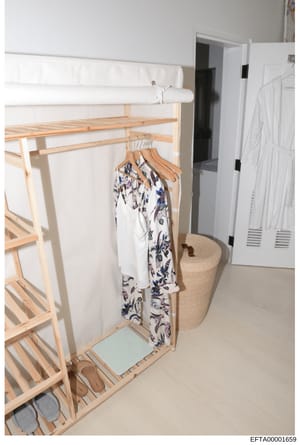 This is a photograph of an interior room space showing a wooden clothing rack with hanging garments, storage furniture, and household items. The image appears to be evidence documentation, likely taken during an investigation of a residence. The room