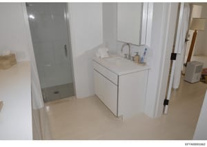 This is a photograph of a modern bathroom interior, likely taken during a property inventory or evidence documentation process. The image shows a clean, contemporary bathroom with white cabinetry, a double vanity sink, glass-enclosed shower, and stan