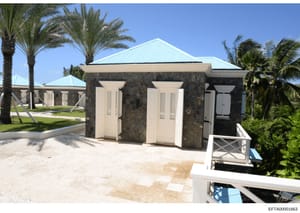 This photograph shows a modern beachfront building with distinctive turquoise metal roofing, dark stone/concrete walls, and cream-colored double doors. The structure is surrounded by palm trees and manicured landscaping, suggesting a high-end tropica
