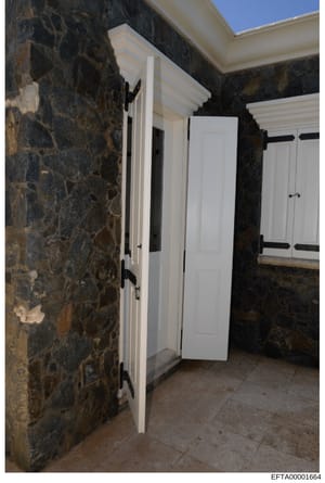 This is a photograph of an outdoor or semi-enclosed courtyard space featuring dark stone walls, white wooden doors with black metal hardware, and tiled flooring. The image appears to be documentation of a property's architectural features and conditi